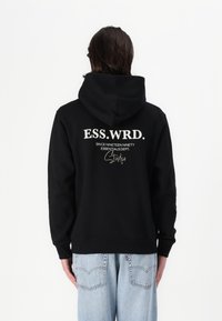 Svart hettegenser med hvit tekst på baksiden: "ESS.WRD. SINCE NINETEEN NINETY ESSENTIALS DEPT. Studio." Justerbar hette, avslappet passform.