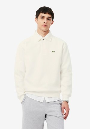 Jonge man die een wit poloshirt met lange mouwen en lichtgrijze broek draagt, staat met één hand in zijn zak tegen een witte achtergrond.