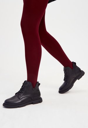 Personne portant des collants opaques rouge foncé et des bottines à lacets noires, debout devant un fond blanc uni.