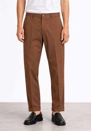 Mężczyzna ubrany w brązowe spodnie z mankietami i zagięciem z przodu oraz czarne wsuwane skórzane buty, stojący na szarej podłodze.