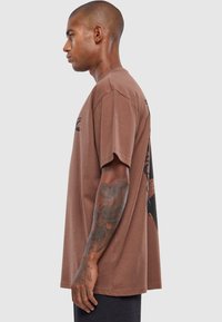 Brun bomulds t-shirt, kortærmet, afslappet pasform, med et grafisk print på ryggen, synlige tatoveringer på armen.