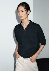 Polo shirt nero a maglia con maniche corte, dotato di due tasche frontali e dettagli in bottoni. Abbinato a pantaloni di colore chiaro.