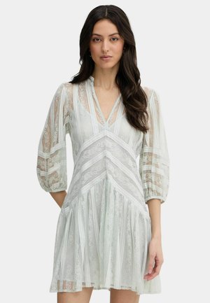 Donna che indossa un vestito bianco in pizzo con scollo a V, maniche a palloncino trasparenti e motivi geometrici in pizzo, in piedi davanti a uno sfondo chiaro e uniforme.