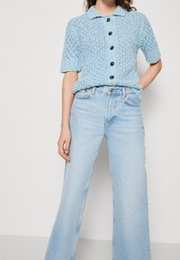 Ljusblå stickad kortärmad tröja med krage och svarta knappar, tillsammans med högmidjade, ljusblå utsvängda jeans.