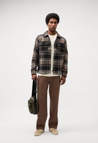 Plaid flannel shirt in brown and black, cream t-shirt, brown wide-leg pants, and beige sneakers. Black strap bag with green accents.