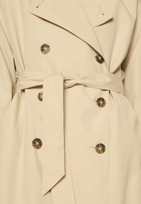 Beige trench coat with double-breasted tortoiseshell buttons and a tied waist belt.