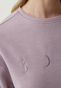 Laventelin värinen huppari, jossa on pehmeä tekstuuri, pyöreä kaula-aukko ja kolme brodeerattua yksityiskohtaa, jotka muistuttavat kuun muotoja edessä.