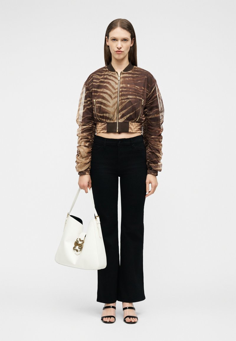 Γυναίκα που φοράει καφέ τζάκετ με ρίγες τίγρης κομμένο στη μέση, μαύρο παντελόνι, μαύρα σανδάλια και κρατάει μια λευκή τσάντα χειρός με χρυσές διακοσμήσεις.