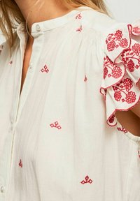 Blusa bianca con ricami floreali rossi, maniche corte arricciate e colletto con bottoni indossata da una persona con capelli chiari.