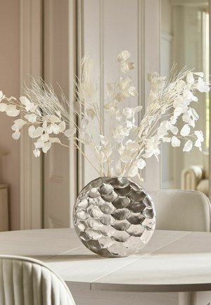 Rund sølvvase med tekstureret mønster, der holder hvide tørrede blomster på et lyst rundt spisebord i et rum med neutrale farver.