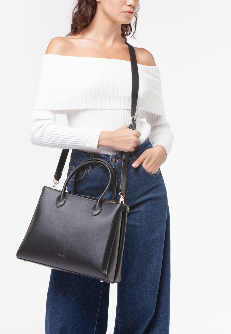 Borsa nera con forma strutturata, texture liscia, con doppio manico superiore, tracolla removibile e dettagli dorati. Indossata sopra una maglietta bianca.