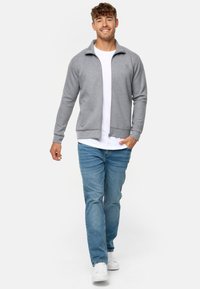 Graue Zip-Jacke mit hohem Kragen und gerippten Bündchen, kombiniert mit einem weißen T-Shirt und hellblauen Jeans. Stehend vor einem einfarbigen Hintergrund.