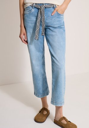 Personne portant un jean court bleu clair avec une ceinture noire et blanche à motifs et des chaussures marron sans lacets. Une main dans la poche, l'autre détendue.