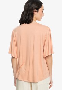 Meruňková tričko s krátkými, volánkovými rukávy a uvolněným střihem. Měkká tkanina, kulatý výstřih a jednoduchý design.