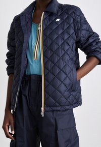 Giacca trapuntata blu navy con colletto rialzato, dotata di zip frontale e dettagli in zip multicolori. Indossata sopra un top teal e pantaloni blu navy.