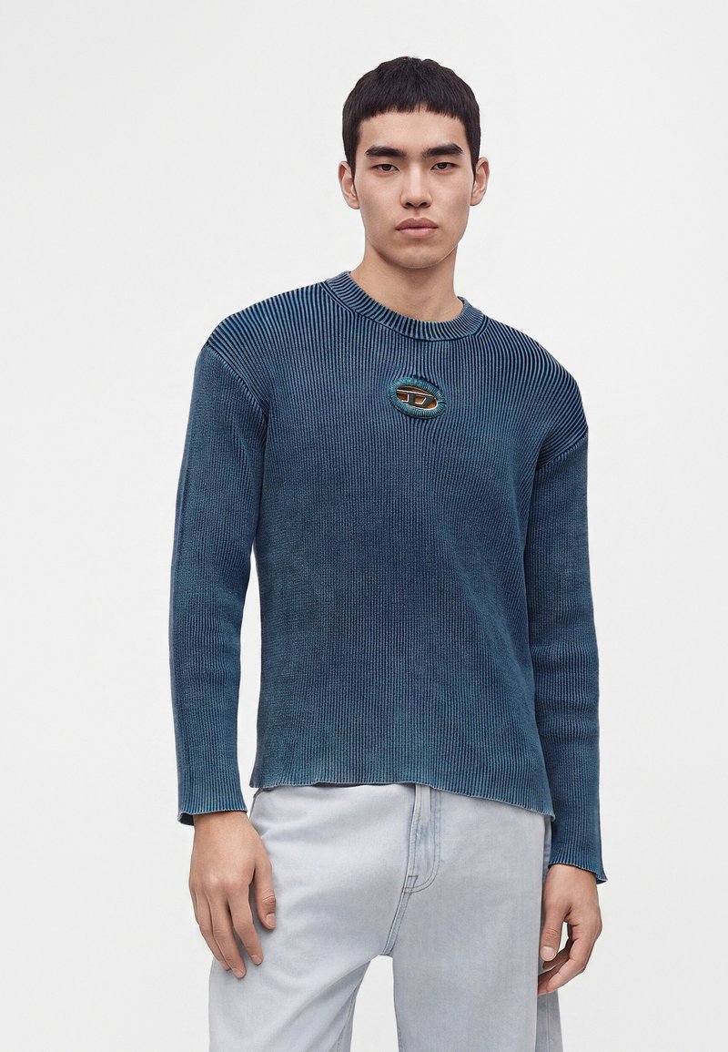Jeune homme aux cheveux courts et noirs portant un pull bleu côtelé avec un petit détail découpé en forme ovale, associé à un pantalon gris clair.