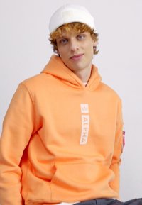 Sudadera naranja con un parche de logo en el frente, capucha con cordones y bolsillo tipo canguro. Llevada con un gorro de punto blanco. Textura suave.