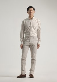 Camisa beige clara de botones, pantalones crema de corte ajustado y zapatos de ante marrón, de pie sobre un fondo neutro. Diseño simple con los botones de la camisa visibles.