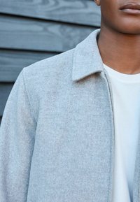 Lichtgrijze zip-jack met een kraag, gemaakt van textuurmateriaal, gelaagd over een wit shirt. Focus op de strakke lijnen van de jack en de zachte stof.