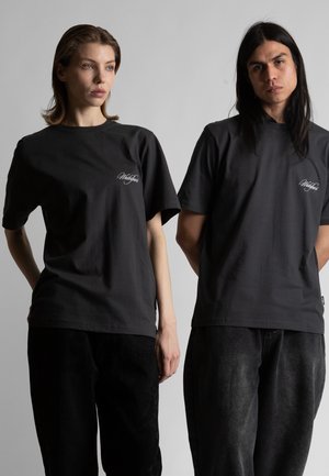 Due modelli indossano t-shirt nere con piccoli loghi bianchi scritti sul lato sinistro del petto, in piedi davanti a uno sfondo grigio chiaro semplice.