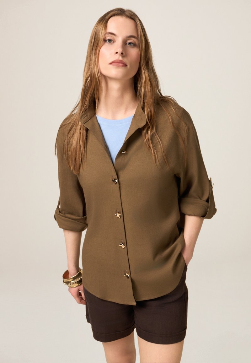 Camicia marrone a bottoni con maniche arrotolate sopra una maglietta azzurro chiaro, abbinata a pantaloni corti marrone scuro con bottoni color oro.
