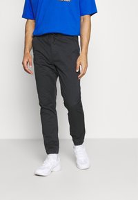 Jogging noirs fuselés avec une taille élastique et des poignets, comportant un cordon de serrage et un tissu léger et lisse. Portés avec des baskets blanches.
