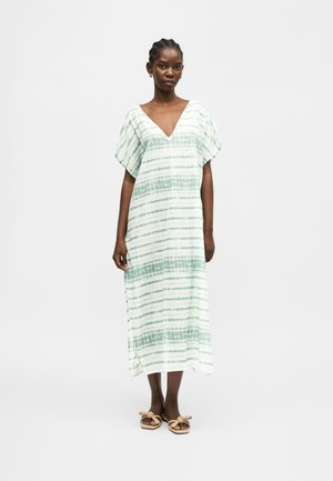 Žena oblečená v voľných bielo-zelených pruhovaných midi šatách s výstrihom do V, krátkymi rukávmi a béžovými sandálmi, stojaca na jednoduchom pozadí.