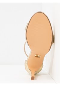 Semelle d'une chaussure à talon beige avec motif texturé, petit talon, et emblème logo doré sur une surface inférieure lisse.