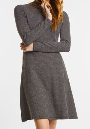 Kobieta w szarej, dzianinowej sukience za kolano z długim rękawem i subtelnym wzorem w kratkę, stojąca z założonymi rękami i dotykająca włosów.
