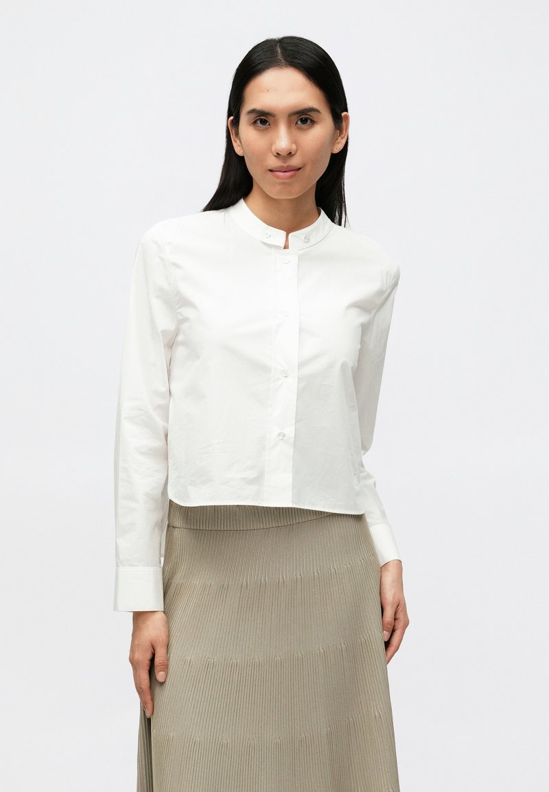 Λευκό κοντό μπλουζάκι με στρογγυλή λαιμόκοψη, μακριά μανίκια και κουμπιά στο μπροστινό μέρος. Συνδυασμένο με ριμπ υφής, μπεζ φούστα μέχρι το γόνατο.