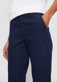 Personne portant un pantalon bleu marine avec la main dans la poche et une chemise blanche rentrée, montrée de la taille au milieu de la cuisse.