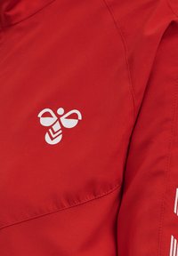 Tissu rouge avec logo d'abeille stylisé blanc et couture détaillée visible sur un vêtement.