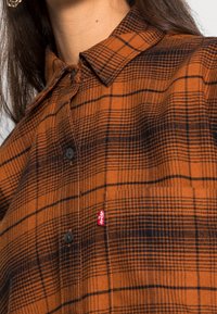 Camicia a quadri marrone e nera con colletto classico, chiusura con bottoni e una piccola etichetta rossa Levi's sul lato sinistro in basso.