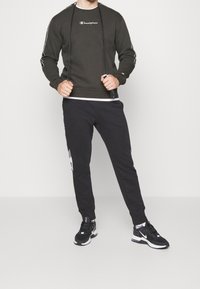 Uomo in felpa Champion nera, pantaloni jogger neri e sneakers Nike nere, che tiene una corda per saltare contro uno sfondo uniforme.
