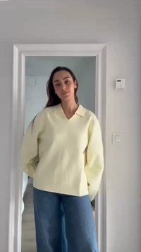 Γυναίκα με μακριά σκούρα μαλλιά, ντυμένη με ανοιχτό κίτρινο πουλόβερ και μπλε τζιν, στέκεται σε ένα κατώφλι με τα χέρια στις τσέπες μπροστά σε έναν απλό τοίχο.