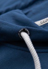 Gros plan sur un tissu en coton bleu avec un cordon blanc passé dans un œillet en métal noir portant l'inscription "ambre wear".