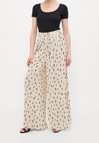 Pantalons larges beiges avec un motif en forme de losange noir, fabriqués en tissu léger avec une texture fluide et taille haute.