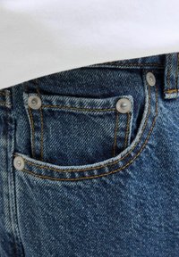 Jeans en denim avec un lavage bleu foncé, présentant des coutures accentuées, des rivets métalliques sur les poches et un petit détail de poche avant.