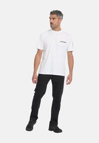 Hombre de pie, con camiseta blanca de manga corta Spyder, pantalones negros y zapatillas deportivas negras, mirando hacia su izquierda sobre un fondo liso.