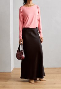Rosa Langarmoberteil kombiniert mit einem schwarzen, bodenlangen Rock. Hält eine karierten rot-schwarzen Handtasche; trägt beige, strukturierte Schuhe.