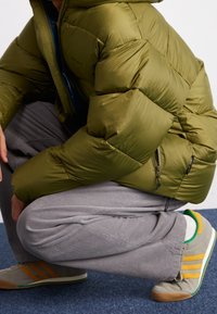 Giacca puffer verde oliva con trapuntatura orizzontale, fodera blu e zip. Pantaloni in velluto grigio e sneakers grigie con dettagli verdi e gialli.