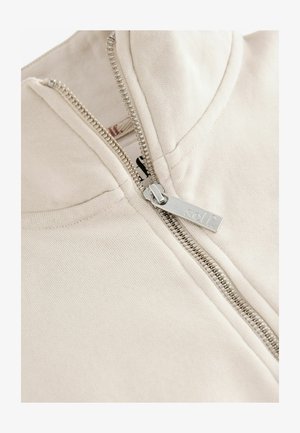 Beige rits-huisvest met een zilveren metalen rits, voorzien van een ribgebreide kraag en een gladde katoenstructuur. Label gedeeltelijk zichtbaar aan de binnenkant.