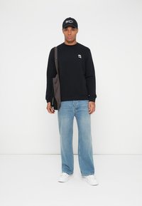 Svarte hettegenser med logo, lyseblå vide jeans, hvite joggesko og svart caps med hvit tekst, bærende en brun skulderveske.