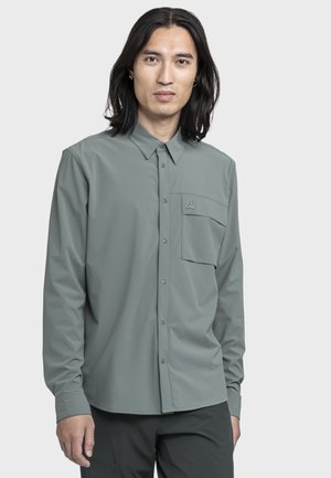 Mann mit langen schwarzen Haaren, der ein graues Hemd mit Knöpfen und Brusttasche trägt, vor einem schlichten hellgrauen Hintergrund stehend.