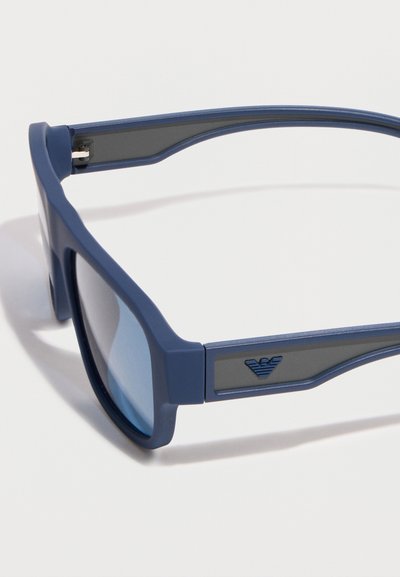 Μπλε ορθογώνια γυαλιά ηλίου με σκουρόχρωμους φακούς και μικρό τρίγωνο λογότυπο με φτερά στο μπράτσο του σκελετού.