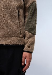 Fleecejacke in Beige mit einem olivgrünen Schulterpanel, ausgestattet mit einer Reißverschlusstasche und einem Logo-Akzent am Ärmel. Strukturierter, gemütlicher Stoff.