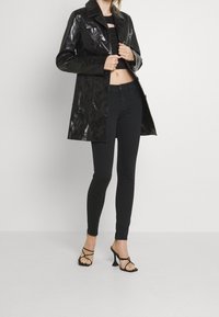 Impermeable negro y brillante con cuello tipo solapa, bolsillos frontales y corte recto. Llevado sobre un top corto negro y jeans ajustados. Sandalias de tacón alto.