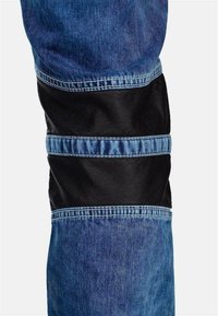 Jeans in denim con un pannello di pelle scuro intorno alla zona del ginocchio, in contrasto con un denim blu più chiaro, con dettagli di cucitura visibili.
