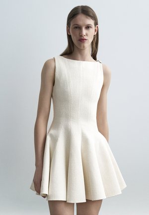 Woman with straight brown hair wearing a sleeveless, textured off-white dress with a flared skirt, standing against a plain light gray background.