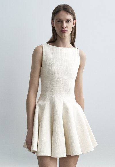 Femme aux cheveux bruns et lisses, vêtue d'une robe sans manches texturée de couleur blanc cassé avec une jupe évasée, debout devant un fond gris clair uni.